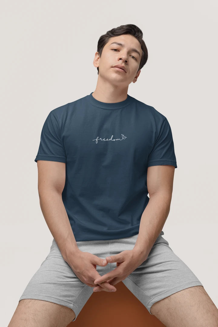 A man wearing a midnight navy t-shirt with a white 'freedom' with bird line art on the chest, paired with light gray shorts, sitting against a light beige background.