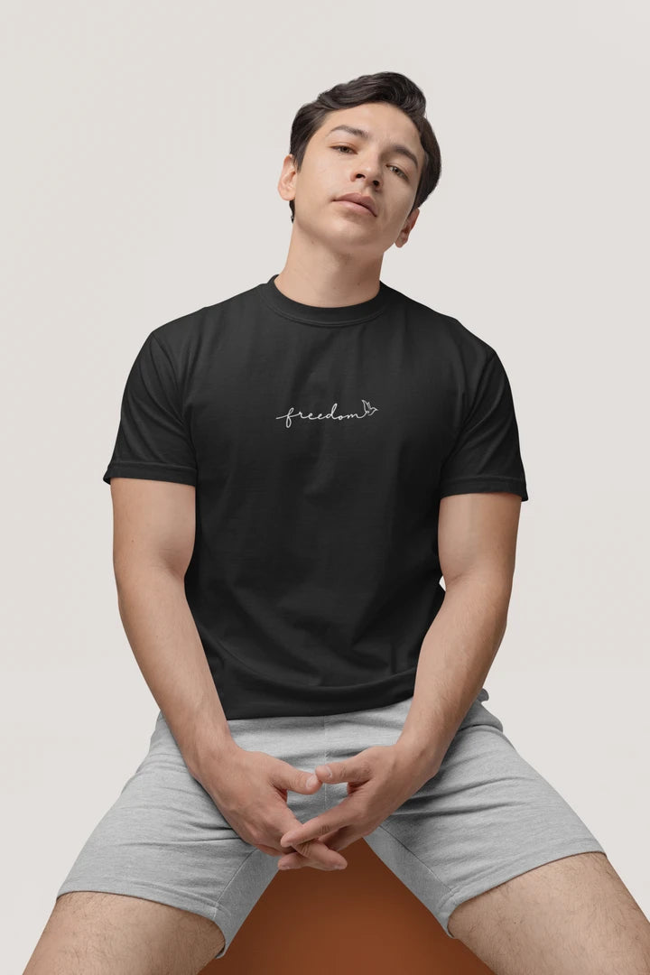 A man wearing a jet black t-shirt with a white 'freedom' graphic with bird line art on the chest, paired with light gray shorts, sitting against a light gray background.