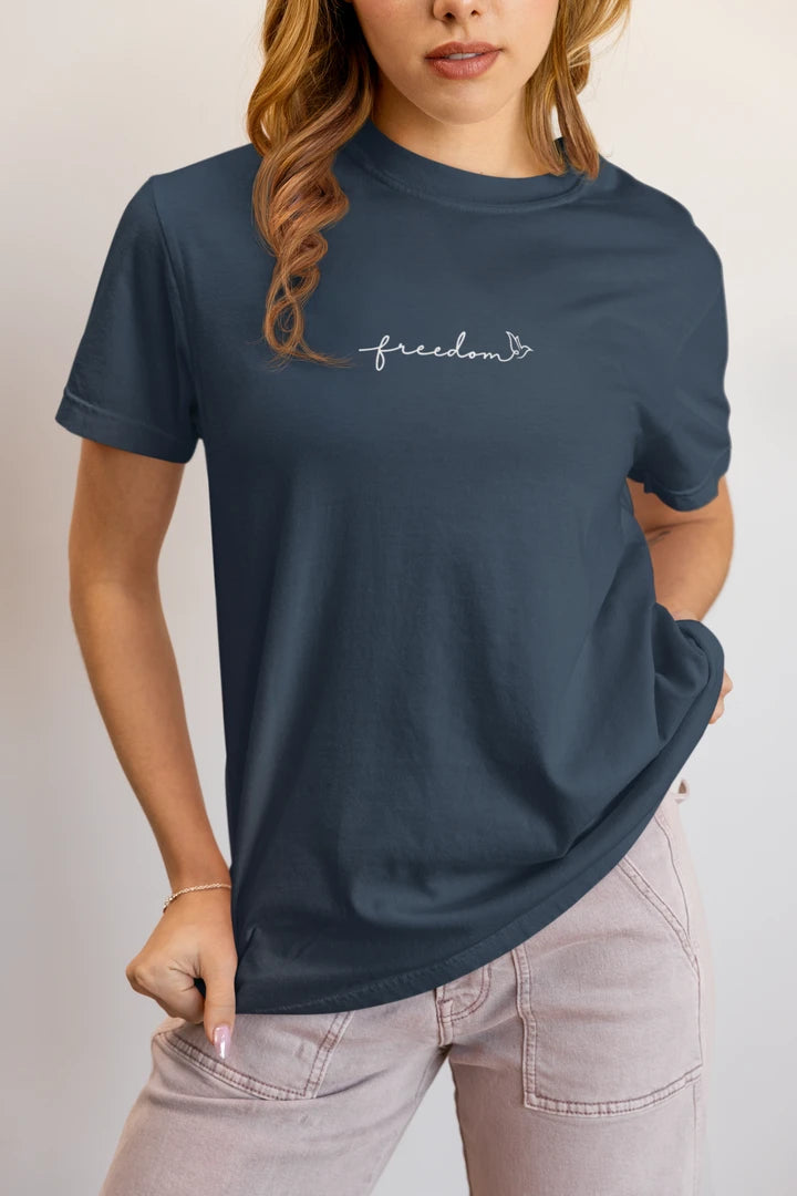 A woman wearing a midnight navy t-shirt with a white 'freedom' graphic with bird line art on the chest, paired with light pink jeans, against a plain light gray background.
