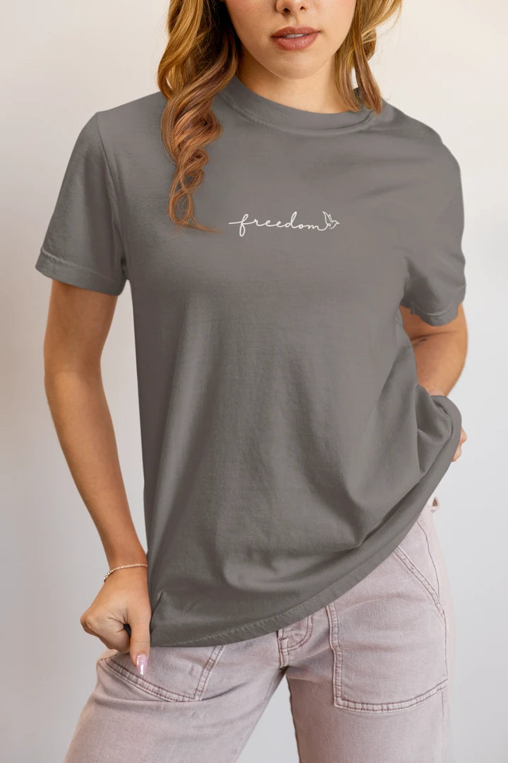 A woman wearing a heather charcoal t-shirt with a white 'freedom' with bird line art on the chest, paired with light pink jeans, against a plain light gray background.