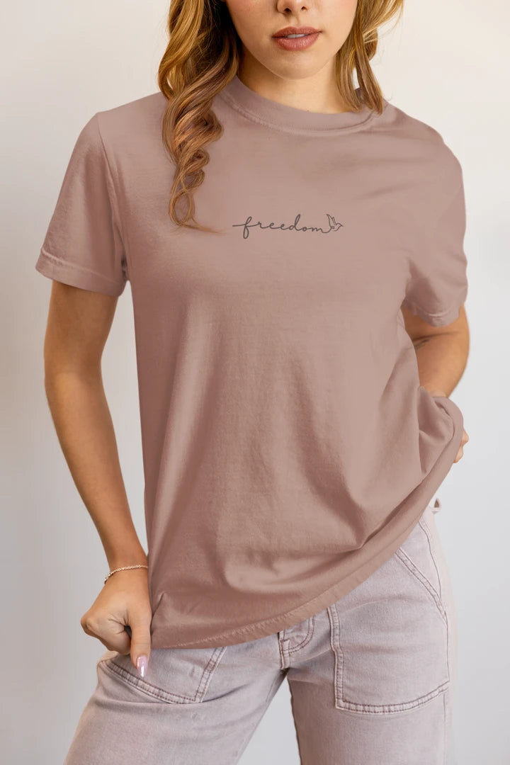 A woman wearing a taupe t-shirt with the word 'freedom' printed with bird line art in black on the chest, paired with light-wash jeans against a plain light gray background.