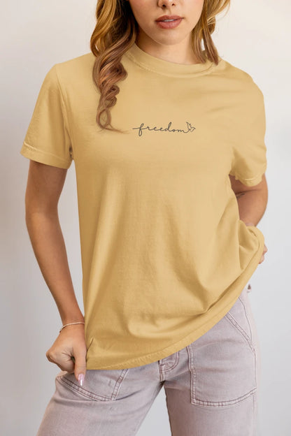 A woman wearing a warm sand t-shirt with 'freedom' printed with bird line art in black on the chest, paired with light gray jeans against a plain light gray background.