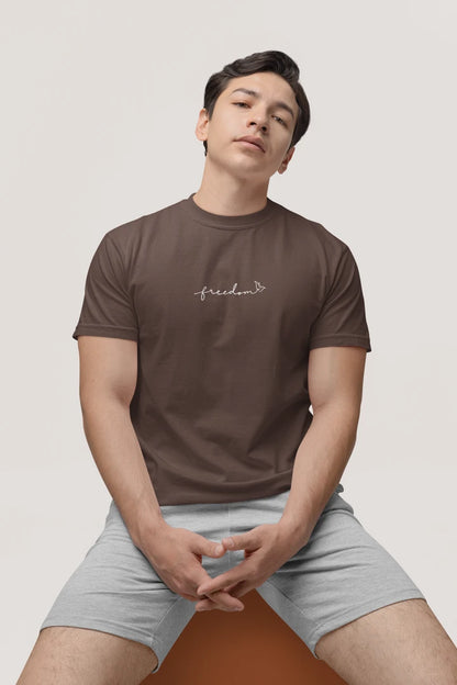 A man wearing a Dark Mocha t-shirt with the text 'Freedom' printed in white with bird line art on the chest, paired with light gray shorts, seated against a light gray background.