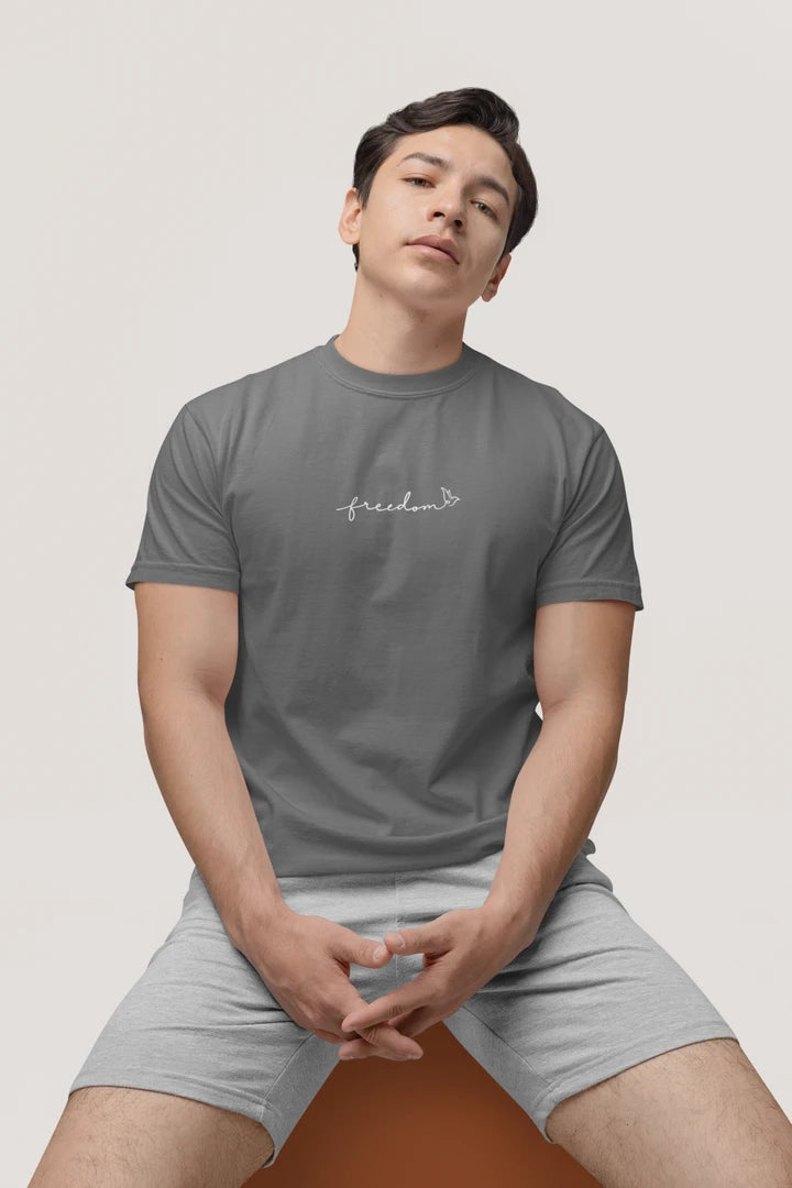 A man wearing a heather charcoal t-shirt with the text 'freedom' printed with bird line art in white on the chest, paired with light gray shorts, seated against a solid light gray background.