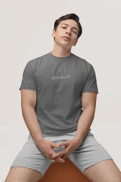 A man wearing a heather charcoal t-shirt with the text 'freedom' printed with bird line art in white on the chest, paired with light gray shorts, seated against a solid light gray background.