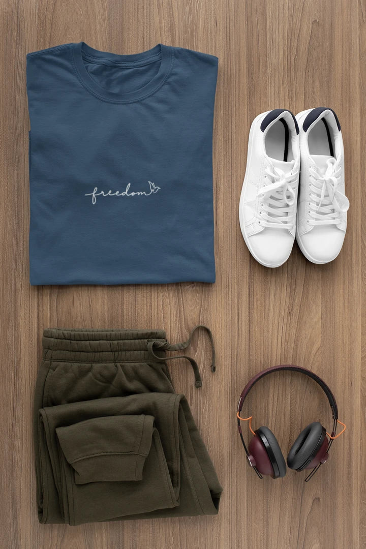 A styled flat-lay of a midnight navy t-shirt with a white 'freedom' graphic with bird line art, olive green drawstring sweatpants, white sneakers, and over-ear headphones on a light brown wood background.