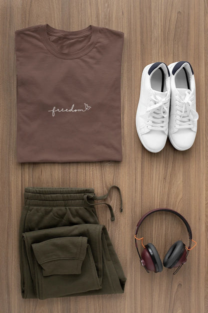 A styled flat-lay outfit on a light brown wood background featuring a Dark Mocha t-shirt with a white 'freedom' logo with bird line art, olive green sweatpants, white sneakers, and black over-ear headphones.