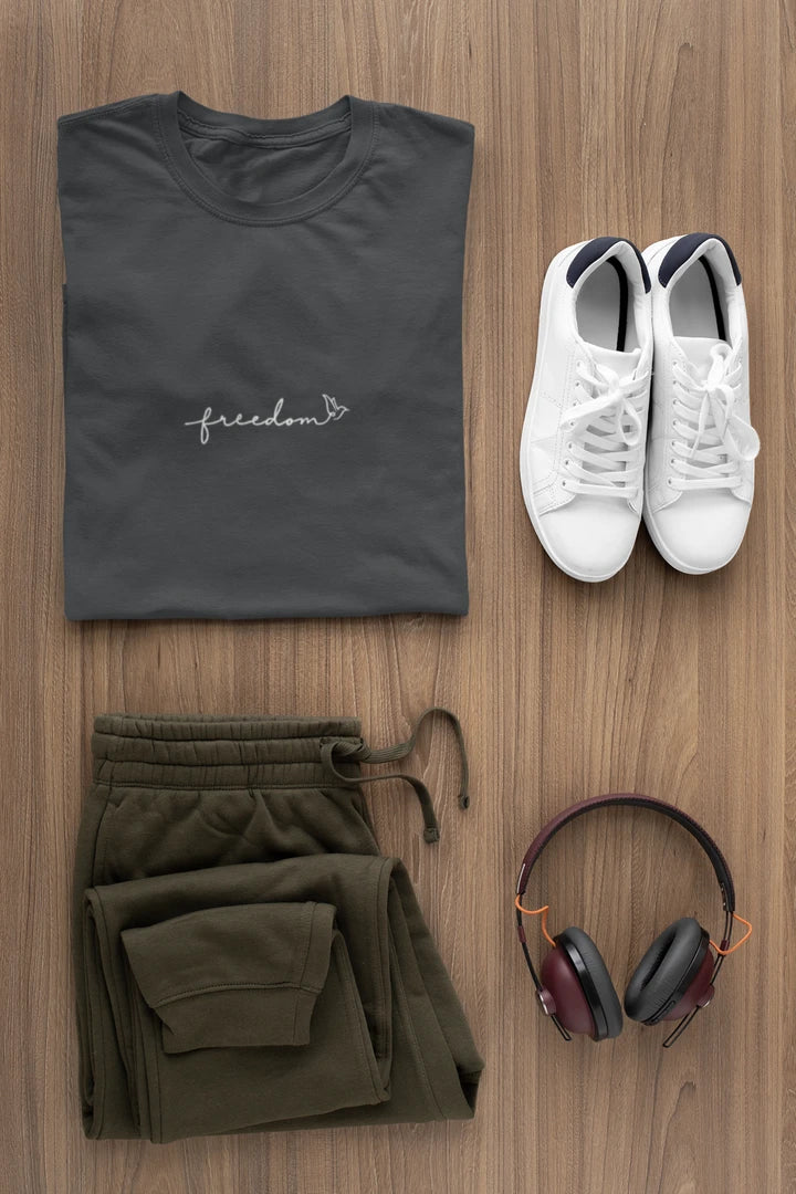 A styled flat-lay outfit on a wood background featuring a graphite t-shirt with a white 'freedom' graphic with bird line art, olive green sweatpants, white sneakers, and black over-ear headphones.