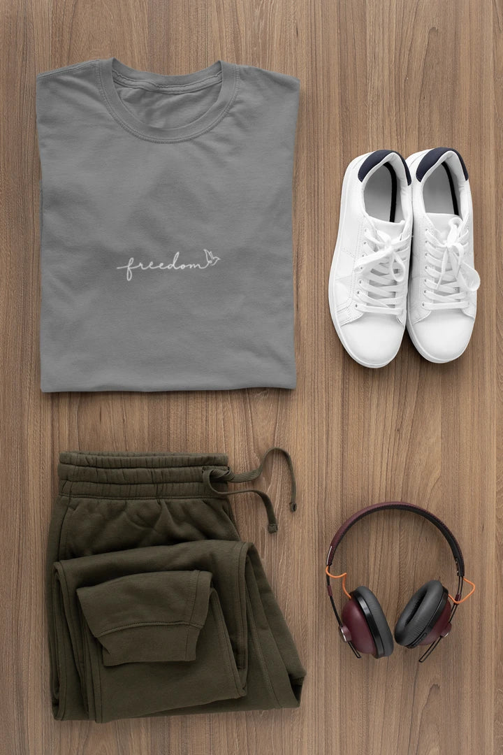 A styled flat-lay of a heather charcoal t-shirt with a white 'freedom' graphic with bird line art, paired with olive green sweatpants, white sneakers, and over-ear headphones on a light brown wood background.