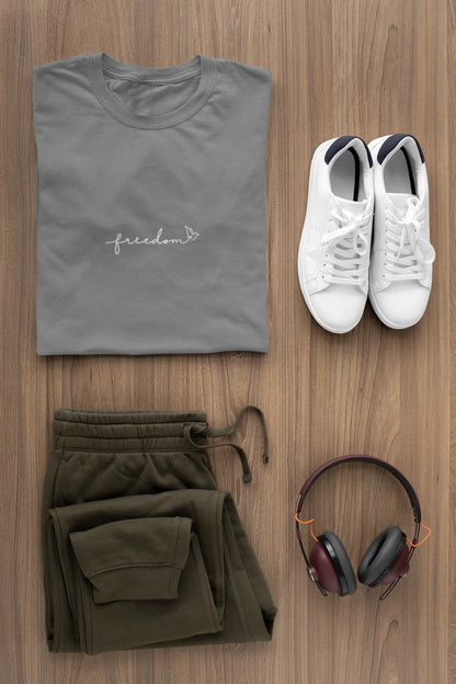 A styled flat-lay of a heather charcoal t-shirt with a white 'freedom' graphic with bird line art, paired with olive green sweatpants, white sneakers, and over-ear headphones on a light brown wood background.