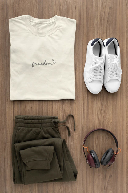 A styled flat-lay of a folded off white t-shirt with 'Freedom' text with bird line art, olive green sweatpants, white sneakers, and over-ear headphones on a light brown wood background.