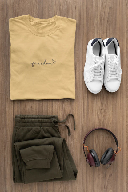 A styled flat-lay of a warm sand t-shirt with 'freedom' printed with bird line art on the chest, paired with olive green sweatpants, white sneakers, and over-ear headphones on a wooden background.