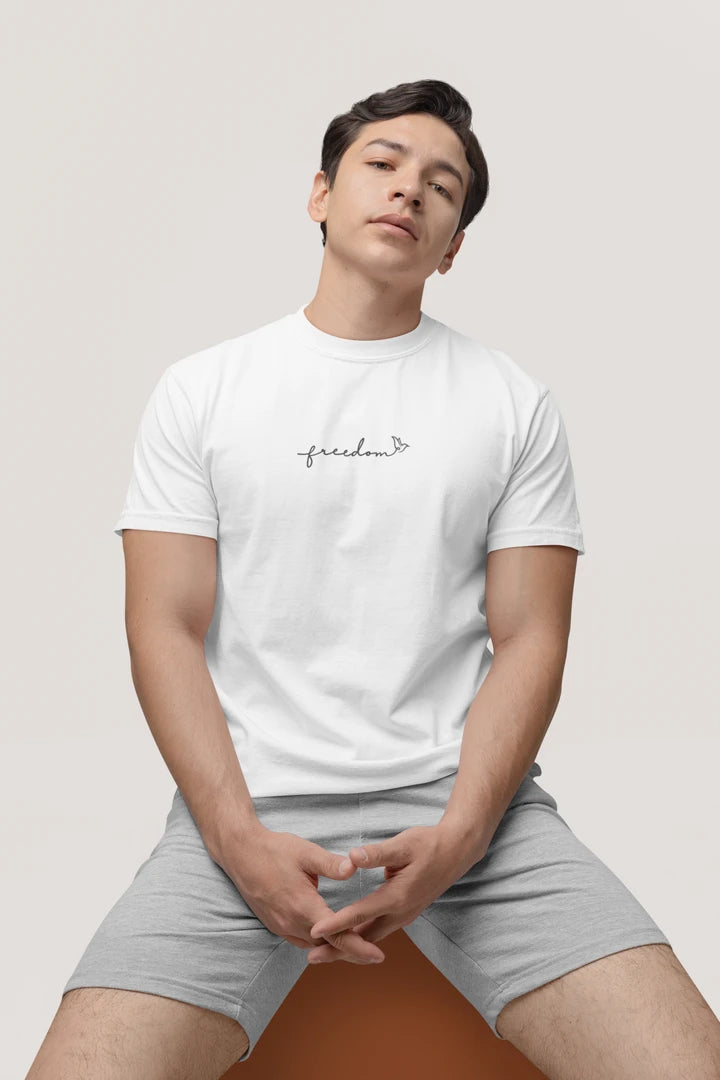 A man wearing a pure white t-shirt with a small black 'freedom' graphic with bird line art on the chest, paired with light gray shorts, sitting against a light gray background.