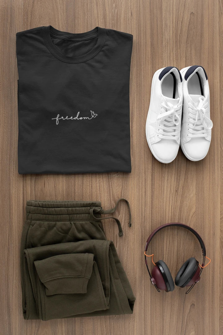A styled flat-lay outfit on a wood background featuring a jet black t-shirt with a white 'freedom' graphic with bird line art, olive green sweatpants, white sneakers, and over-ear headphones.