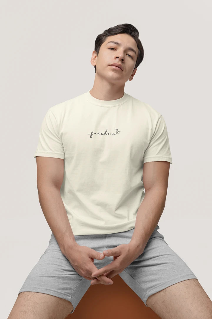 A man wearing a off white t-shirt with a black 'freedom' graphic with bird line art on the chest, paired with light gray shorts, sitting against a solid light gray background.
