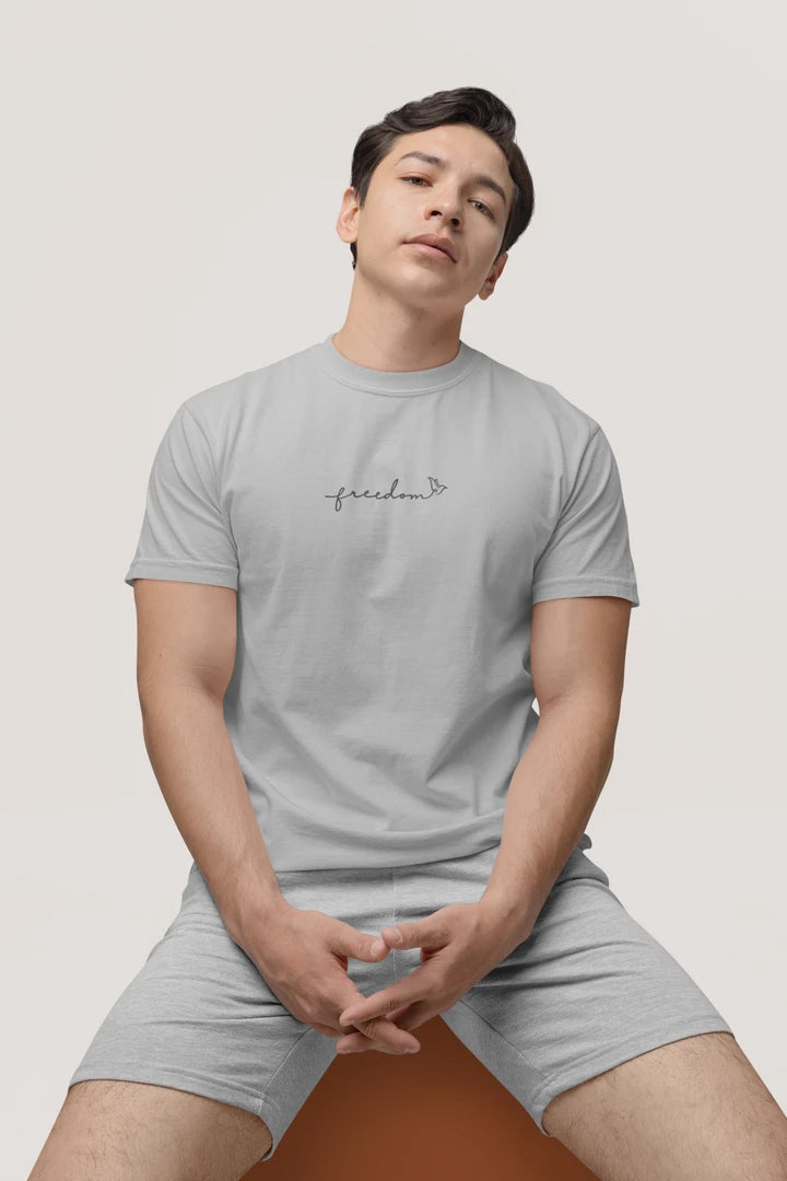 A man wearing a heather grey t-shirt with a black 'freedom' graphic with bird line art on the chest, paired with matching gray shorts, sitting against a light gray background.