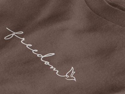Close-up of a dark mocha crewneck t-shirt featuring the word 'freedom' embroidered in white script across the chest, accompanied by a small white bird silhouette.