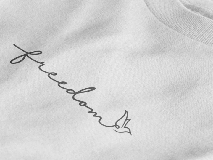 Close-up of a pure white crewneck t-shirt featuring a black cursive script graphic that reads 'freedom' with a dove design at the end.