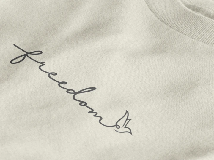 Close-up of a off white crewneck t-shirt featuring the word 'freedom' in black cursive script with a small bird outline, set against a clean white background.