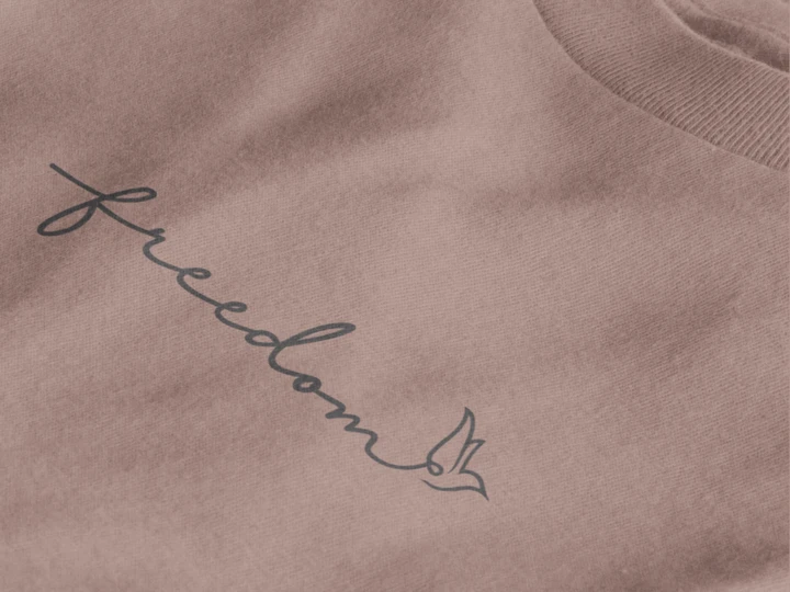 Close-up of a taupe crewneck sweatshirt featuring the word 'freedom' in black cursive script with a small bird silhouette, set against a clean white background.