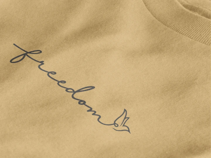 Close-up of a warm sand crewneck sweatshirt featuring the word 'freedom' in dark gray cursive script with a small bird silhouette at the end.