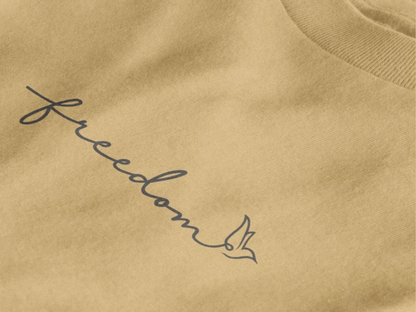 Close-up of a warm sand crewneck sweatshirt featuring the word 'freedom' in dark gray cursive script with a small bird silhouette at the end.