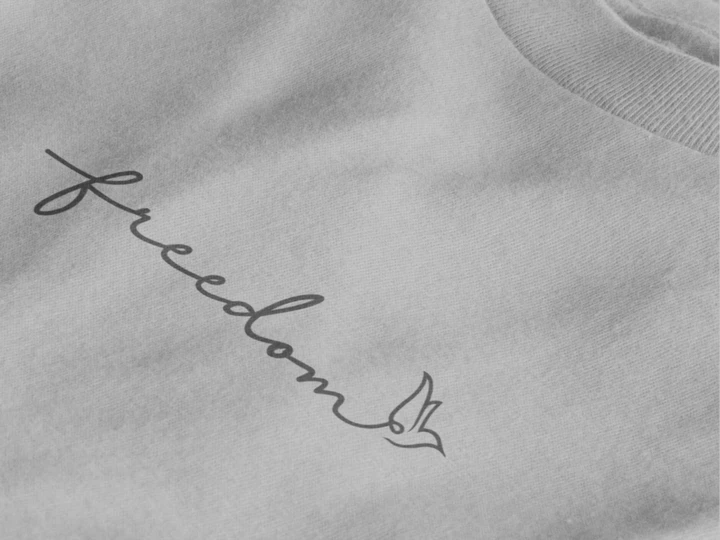 Close-up of a heather grey crewneck sweatshirt featuring the word 'freedom' in black cursive script with a small bird graphic at the end.