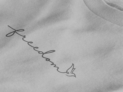 Close-up of a heather grey crewneck sweatshirt featuring the word 'freedom' in black cursive script with a small bird graphic at the end.
