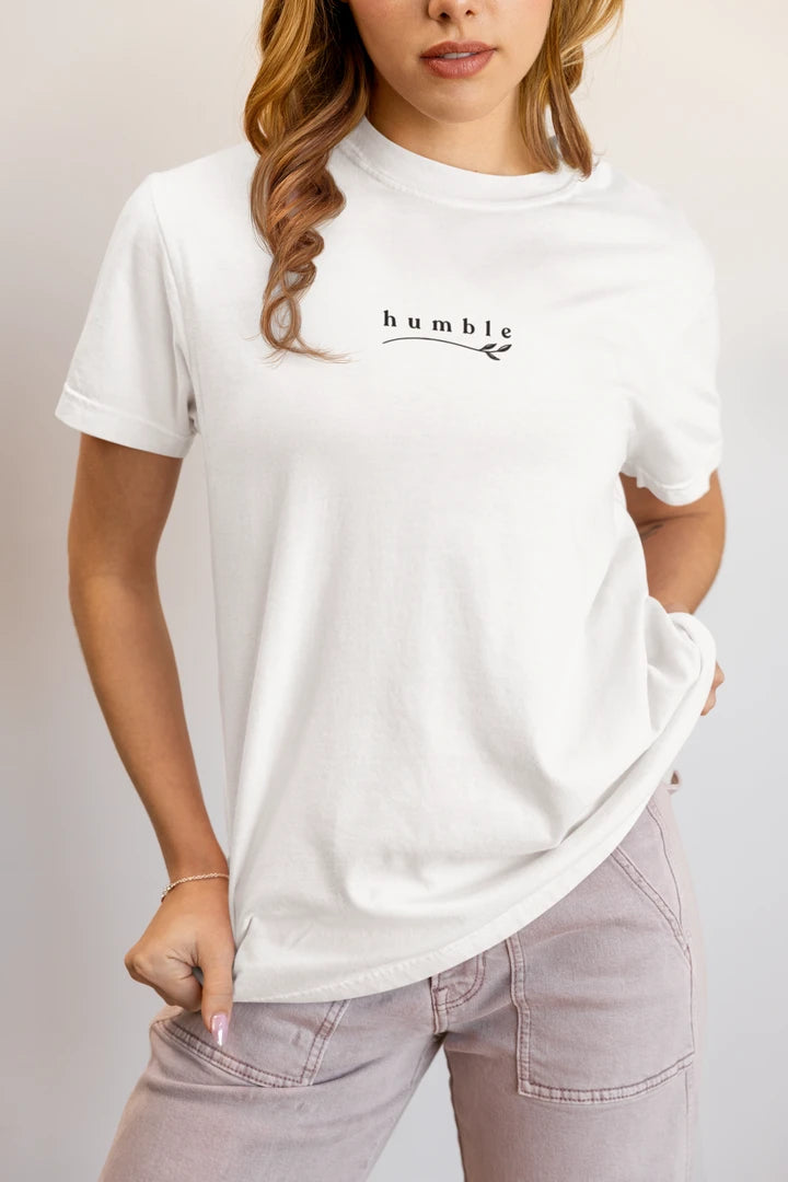 A woman wearing a pure white short-sleeve t-shirt with a black 'humble' graphic on the chest, paired with light gray jeans, against a plain light gray background.