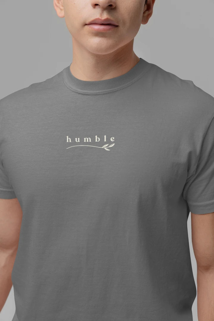 A person wearing a heather charcoal t-shirt with a small white graphic on the chest that reads 'humble' with a leaf design underneath, set against a solid gray background.