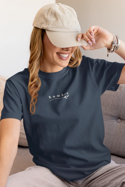 A woman wearing a midnight navy t-shirt with a white 'humble' graphic on the chest, paired with a beige baseball cap and light gray pants, sitting on a gray couch.