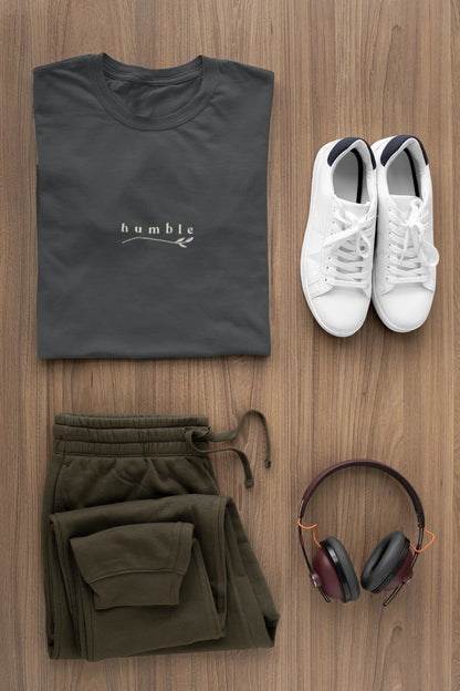 A styled flat-lay outfit on a wood background featuring a Graphite t-shirt with a white 'humble' logo, olive green sweatpants, white sneakers, and black over-ear headphones.