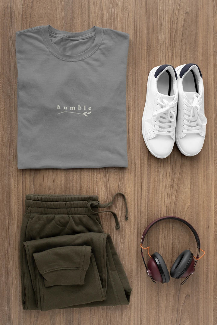 A styled flat-lay of a heather charcoal t-shirt with a white 'humble' logo, paired with olive green sweatpants, white sneakers, and over-ear headphones on a light brown wood background.