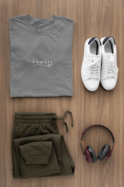 A styled flat-lay of a heather charcoal t-shirt with a white 'humble' logo, paired with olive green sweatpants, white sneakers, and over-ear headphones on a light brown wood background.