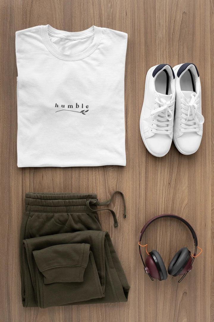 A styled flat-lay outfit on a wood floor featuring a pure white t-shirt with a black 'humble' logo, olive green sweatpants, white sneakers, and over-ear headphones.