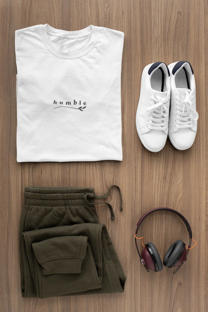 A styled flat-lay outfit on a wood floor featuring a pure white t-shirt with a black 'humble' logo, olive green sweatpants, white sneakers, and over-ear headphones.