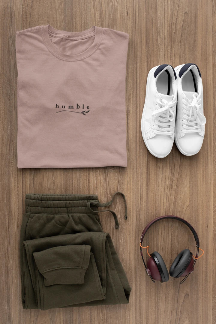 A styled flat-lay outfit on a wood floor featuring a folded taupe t-shirt with the 'humble' logo, olive green sweatpants, white sneakers, and over-ear headphones.