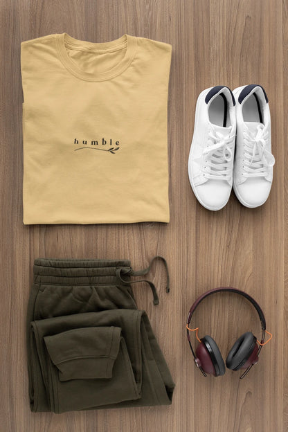 A styled flat-lay outfit on a wood background featuring a folded warm sand t-shirt with 'humble' printed on the chest, olive green sweatpants, white sneakers, and over-ear headphones.