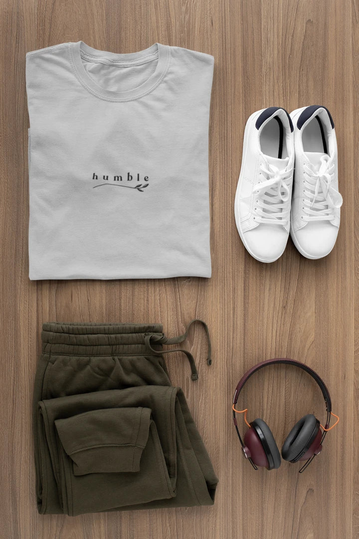 A styled flat-lay of a heather grey t-shirt with 'humble' printed on the chest, paired with olive green drawstring sweatpants, white sneakers, and over-ear headphones on a wooden background.