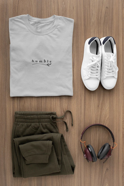 A styled flat-lay of a heather grey t-shirt with 'humble' printed on the chest, paired with olive green drawstring sweatpants, white sneakers, and over-ear headphones on a wooden background.