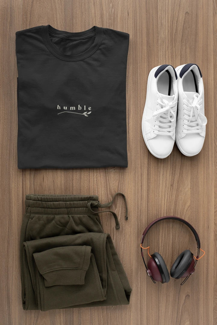 A styled flat-lay outfit on a wood background featuring a jet black t-shirt with a white 'humble' logo, olive green sweatpants, white sneakers, and over-ear headphones.
