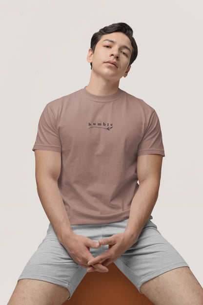 A man wearing a taupe t-shirt with a small black 'humble' graphic on the chest, paired with light gray shorts, sitting against a light gray background.