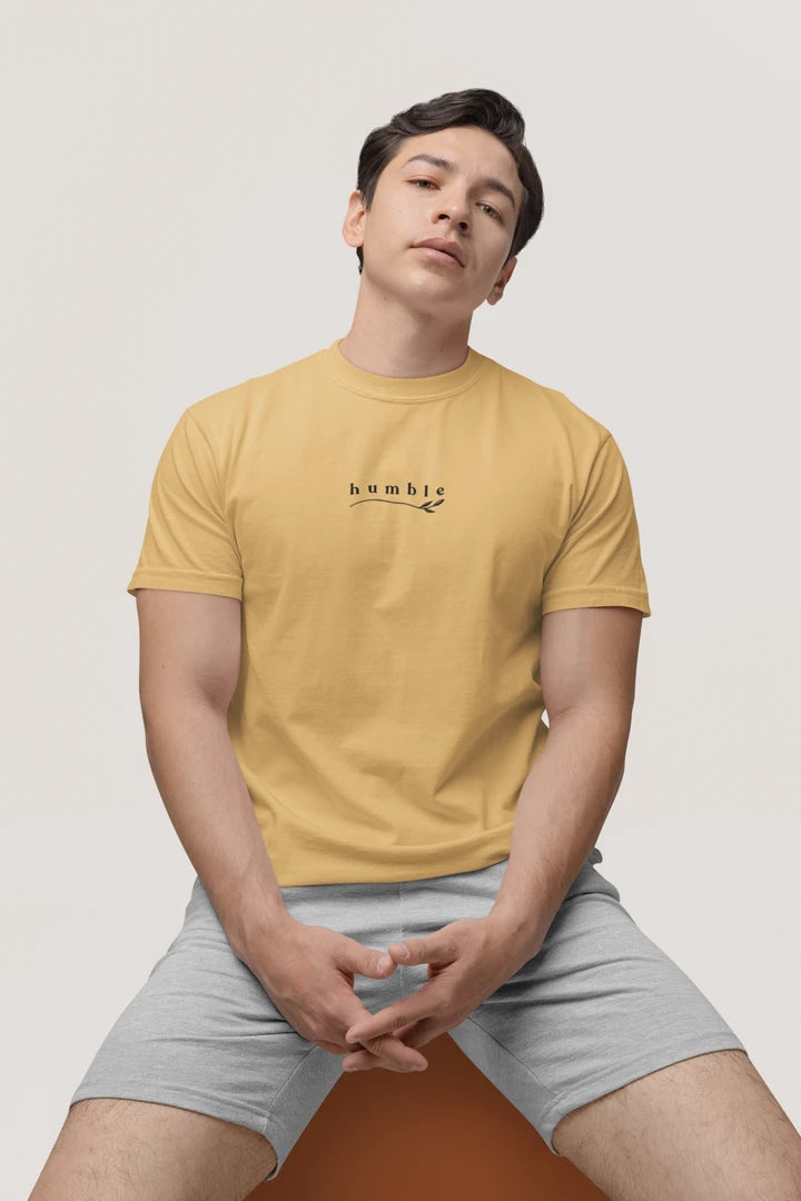 A man wearing a warm sand t-shirt with a black 'humble' graphic on the chest, paired with light gray shorts, sitting against a light gray background.