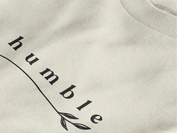 Close-up of a off white crewneck t-shirt featuring a black graphic print that reads 'humble' in a stylized font with a leaf motif.