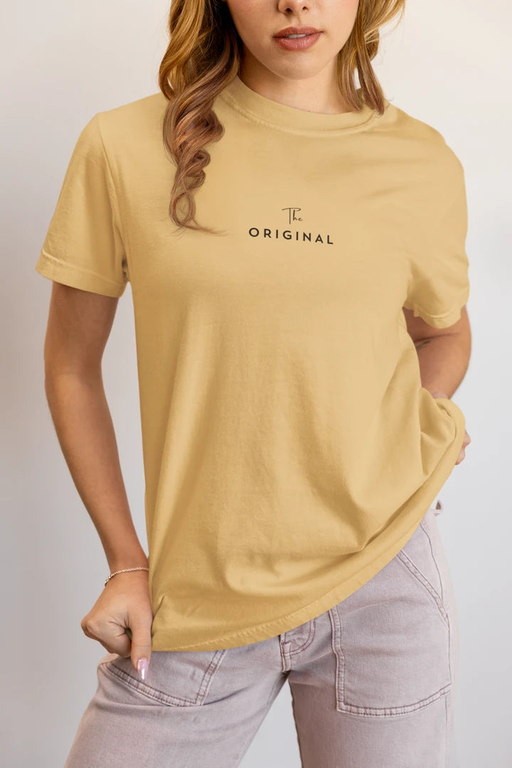 A woman wearing a warm sand t-shirt with 'THE ORIGINAL' printed in black on the chest, paired with light gray jeans against a plain light gray background.