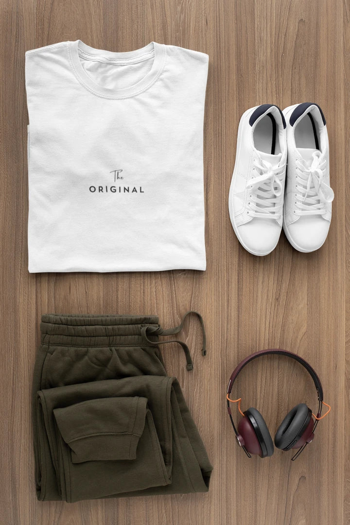 A styled flat-lay outfit on a wood floor featuring a Pure white t-shirt with 'The ORIGINAL' text, olive green sweatpants, white sneakers, and over-ear headphones.
