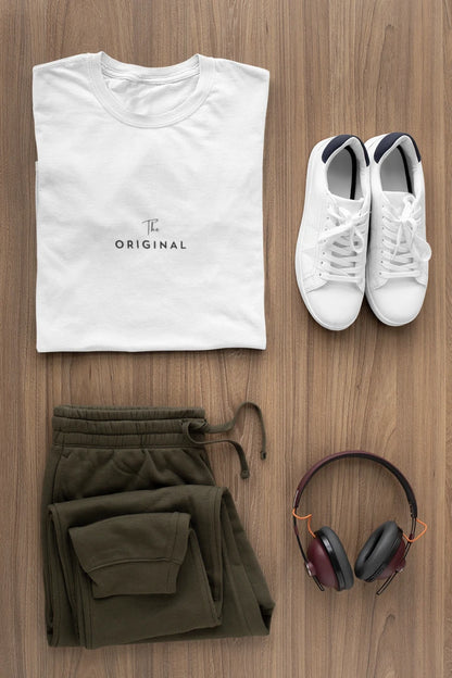 A styled flat-lay outfit on a wood floor featuring a Pure white t-shirt with 'The ORIGINAL' text, olive green sweatpants, white sneakers, and over-ear headphones.