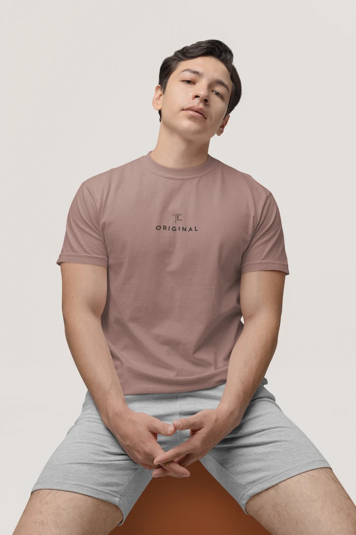 A man wearing a taupe t-shirt with a small black 'The Original' graphic on the chest, paired with light gray shorts, sitting against a light gray background.