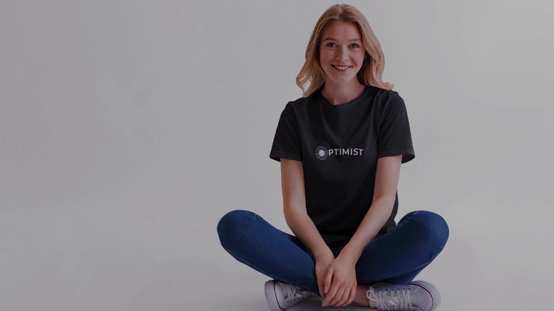 Person wearing a black t-shirt with 'Optimist' logo sitting cross-legged on a white background