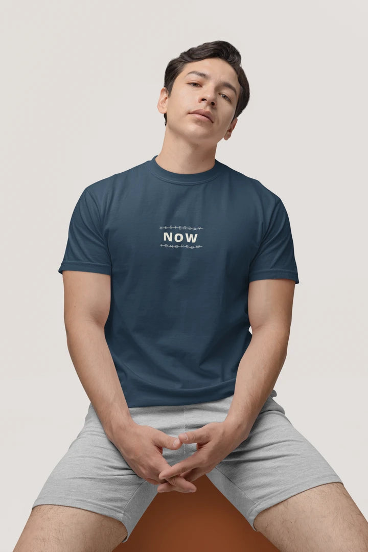 A man wearing a Midnight Navy crewneck t-shirt with a white 'YESTERDAY NOW Tomorrow' logo on the chest, paired with light gray shorts, sitting against a light beige background.